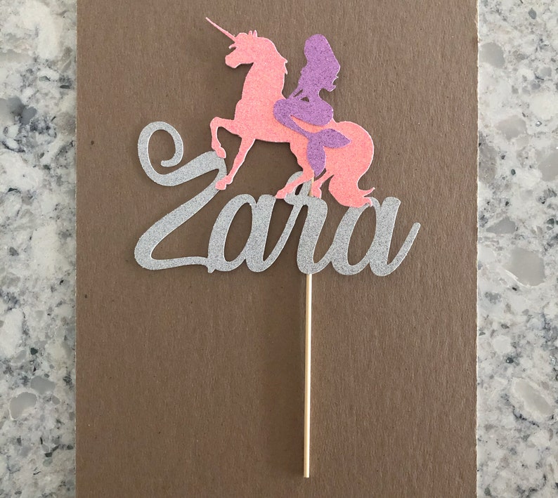 May include: A silver glitter cake topper with the name "Zara" written in cursive. A pink unicorn with a purple mermaid riding on its back.