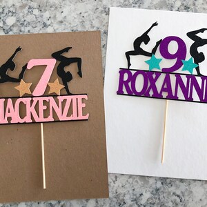 Puede incluir: Dos adornos para pasteles de color rosa y morado con siluetas negras de gimnastas. El adorno rosa tiene el n&uacute;mero 7 y el nombre Mackenzie. El adorno morado tiene el n&uacute;mero 9 y el nombre Roxanne.