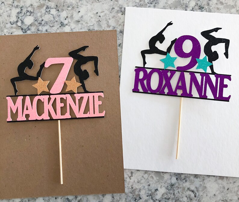 Puede incluir: Dos adornos para pasteles de color rosa y morado con siluetas negras de gimnastas. El adorno rosa tiene el n&uacute;mero 7 y el nombre Mackenzie. El adorno morado tiene el n&uacute;mero 9 y el nombre Roxanne.
