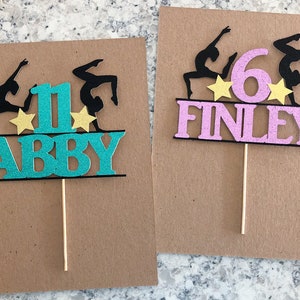 Puede incluir: Dos adornos para pasteles con una silueta de gimnasta, un n&uacute;mero y un nombre. El primer adorno es turquesa con el n&uacute;mero 1 y el nombre Abby. El segundo adorno es rosa con el n&uacute;mero 6 y el nombre Finley.