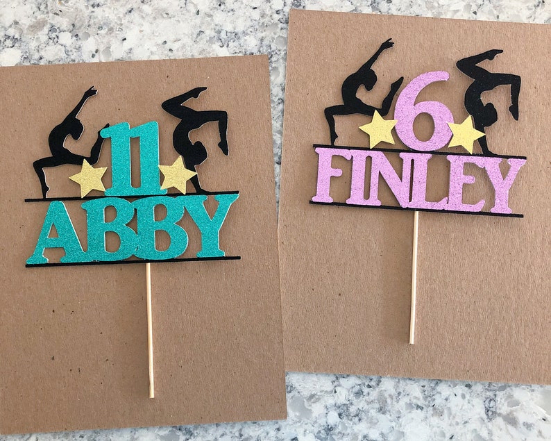 Puede incluir: Dos adornos para pasteles con una silueta de gimnasta, un n&uacute;mero y un nombre. El primer adorno es turquesa con el n&uacute;mero 1 y el nombre Abby. El segundo adorno es rosa con el n&uacute;mero 6 y el nombre Finley.