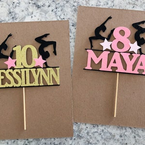 Puede incluir: Dos adornos para pasteles con siluetas negras de gimnastas con estrellas rosas y un n&uacute;mero dorado o rosa. Los adornos tambi&eacute;n incluyen los nombres "Jessilynn" y "Maya".
