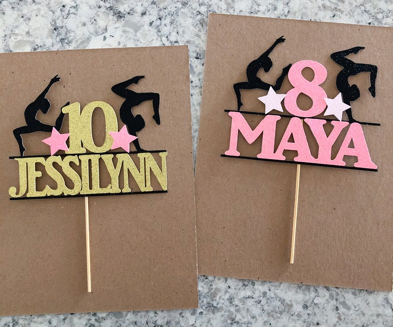 Puede incluir: Dos adornos para pasteles con siluetas negras de gimnastas con estrellas rosas y un n&uacute;mero dorado o rosa. Los adornos tambi&eacute;n incluyen los nombres "Jessilynn" y "Maya".