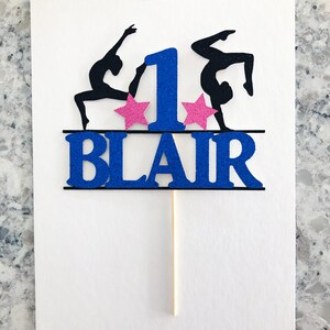 Puede incluir: Topper para pastel de purpurina azul y negro con dos gimnastas, una estrella rosa a cada lado del n&uacute;mero uno y el nombre "BLAIR" debajo.