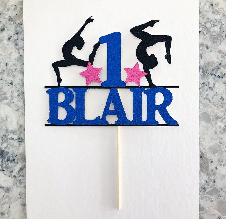 Puede incluir: Topper para pastel de purpurina azul y negro con dos gimnastas, una estrella rosa a cada lado del n&uacute;mero uno y el nombre "BLAIR" debajo.