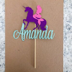 May include: A teal glitter cake topper with the name "Amanda" and a purple unicorn with a pink mermaid riding on its back.