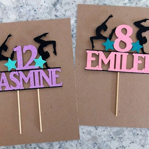 Puede incluir: Dos adornos para pasteles de color rosa y negro con siluetas de gimnastas, estrellas y un n&uacute;mero. El primer adorno dice "12 JASMINE" y el segundo dice "8 EMILIE".