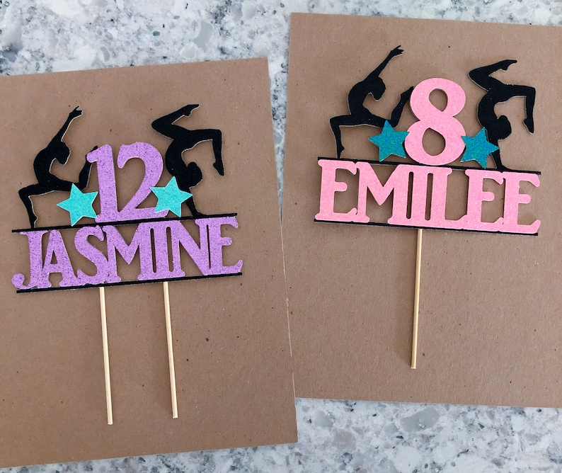 Puede incluir: Dos adornos para pasteles de color rosa y negro con siluetas de gimnastas, estrellas y un n&uacute;mero. El primer adorno dice "12 JASMINE" y el segundo dice "8 EMILIE".