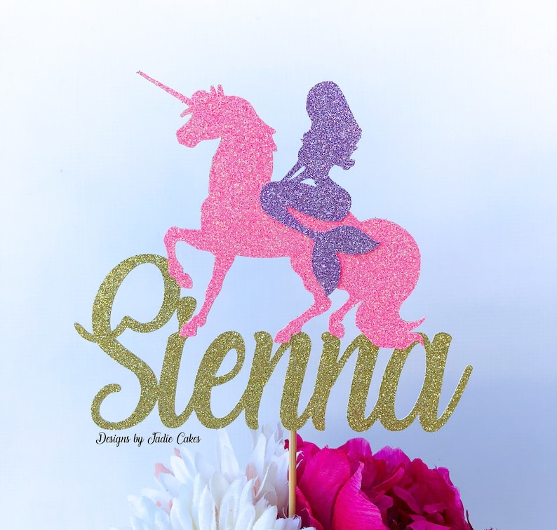 May include: A pink glitter unicorn with a purple glitter mermaid riding on its back. The name "Sienna" is written in gold glitter below the unicorn. The cake topper is on a wooden stick.