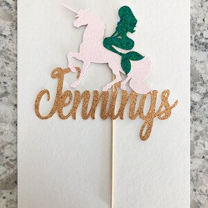 May include: Gold glitter cake topper with the name "Jennings" written in cursive. A pink glitter unicorn with a green glitter mermaid riding on its back.