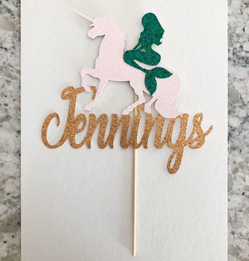 May include: Gold glitter cake topper with the name "Jennings" written in cursive. A pink glitter unicorn with a green glitter mermaid riding on its back.