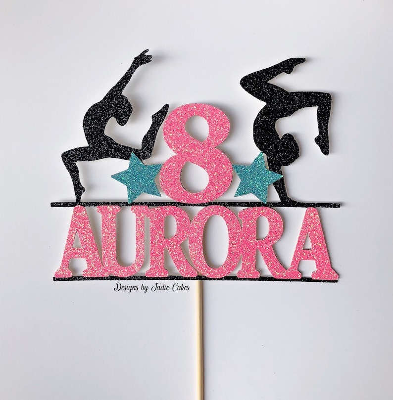 Puede incluir: Topper de pastel de purpurina rosa y negra con dos siluetas de gimnastas, un n&uacute;mero 8 rosa y el nombre "AURORA" en purpurina rosa.