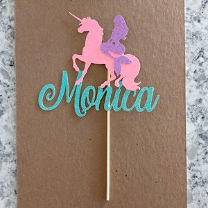 May include: A teal glitter cake topper with the name "Monica" and a pink glitter unicorn with a purple mermaid riding on its back.