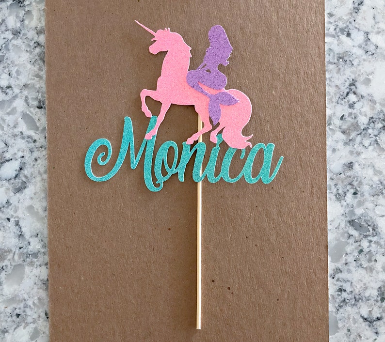 May include: A teal glitter cake topper with the name "Monica" and a pink glitter unicorn with a purple mermaid riding on its back.