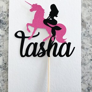 May include: Pink unicorn cake topper with a black mermaid silhouette riding on its back. The topper has the name "Tasha" written in black cursive.