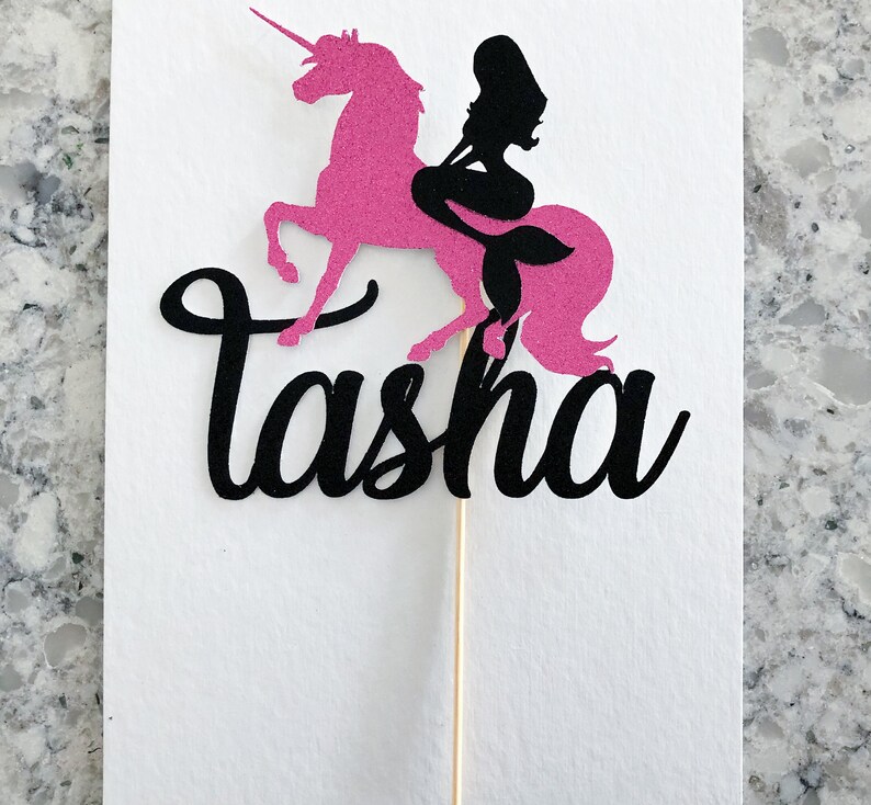 May include: Pink unicorn cake topper with a black mermaid silhouette riding on its back. The topper has the name "Tasha" written in black cursive.
