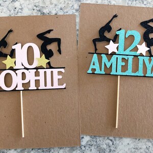 Puede incluir: Dos adornos para pasteles con siluetas de gimnastas en posici&oacute;n de espalda arqueada. Los adornos est&aacute;n decorados con estrellas doradas y los nombres "Sophie" y "Ameilya" con sus respectivas edades, 10 y 12.