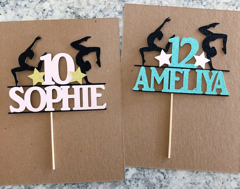 Puede incluir: Dos adornos para pasteles con siluetas de gimnastas en posici&oacute;n de espalda arqueada. Los adornos est&aacute;n decorados con estrellas doradas y los nombres "Sophie" y "Ameilya" con sus respectivas edades, 10 y 12.