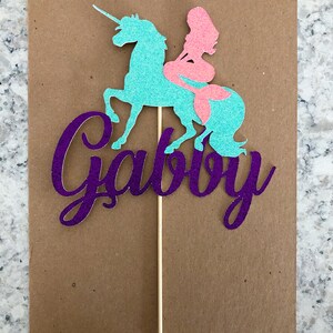 May include: Purple glitter cake topper with the name "Gabby" written in cursive. A teal unicorn with a pink mermaid riding on its back.