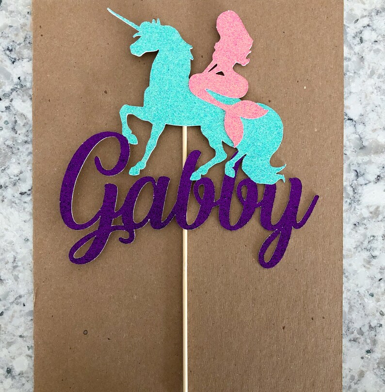 May include: Purple glitter cake topper with the name "Gabby" written in cursive. A teal unicorn with a pink mermaid riding on its back.