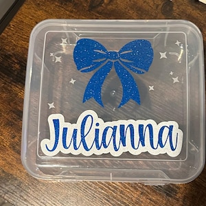 May include: A clear plastic storage box with a blue glitter bow and the name "Julianna" written in white glitter.