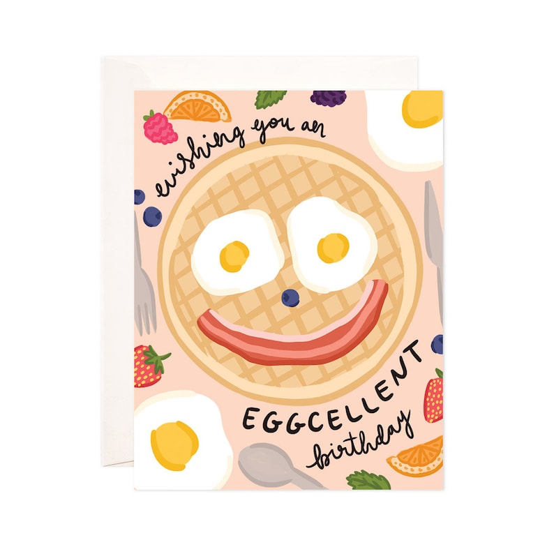 May include: A pink birthday card with a waffle, eggs, and bacon illustration. The waffle has a smiley face and the text "Wishing you an eggcellent birthday" is written on the card.