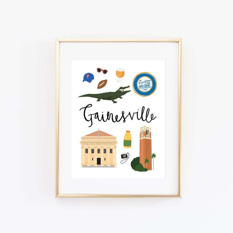 May include: A white framed print with a gold frame. The print features a variety of illustrations representing Gainesville, Florida, including an alligator, a building, a tower, a football, sunglasses, a beer, and a city seal. The text "Gainesville" is written in black cursive.