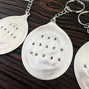 May include: Three silver spoon keychains with different engraved text. The first keychain says "THE CABIN", the second keychain says "THE BEACH HOUSE", and the third keychain says "THE RIVER HOUSE".