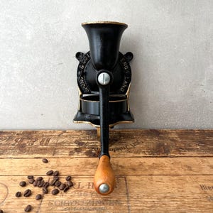 Op de afbeelding: Een witte keramische mok met een zwarte handgreep en de tekst "But First, Coffee" in een vetgedrukte, zwarte letter. De mok staat tegen een vage achtergrond, wat een gezellige setting suggereert.