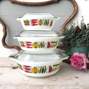 May include: Three white Pyrex bowls with glass lids, stacked on top of each other. The bowls are decorated with a colourful vegetable pattern, including radishes, onions, peas, and tomatoes. The bowls are on a light surface.