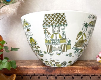 May include: A large white ceramic bowl with a green and brown pattern of people and buildings. The bowl has a wide rim and a rounded bottom.