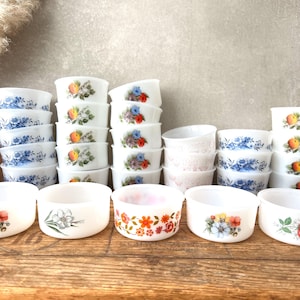 Puede incluir: Una colección de cuencos de cerámica blancos de estilo vintage con diseños florales y frutales. Los cuencos presentan varios patrones, incluyendo flores azules, melocotones y arreglos florales coloridos. Los cuencos están apilados y dispuestos sobre una superficie de madera.