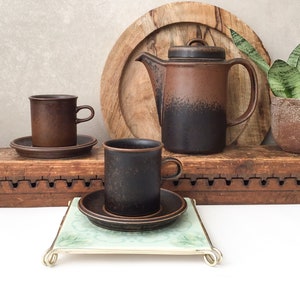 Pode incluir: Um conjunto de canecas de café de cerâmica marrom e preta e um bule de chá combinando com uma bandeja de madeira. As canecas e o bule de chá têm um esmalte salpicado.