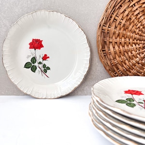 May include: A set of white ceramic plates with a gold rim and a red rose design. The plates are stacked on top of each other.
