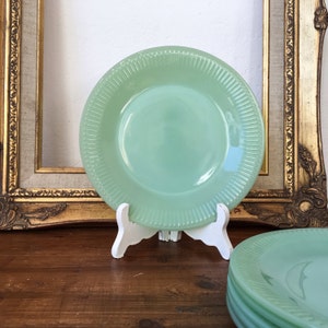 Puede incluir: Un juego de cuatro platos vintage de vidrio verde con borde acanalado. Los platos están apilados en un soporte blanco y están sobre una superficie de madera.