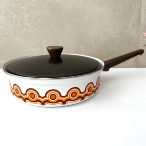 Puede incluir: Sartén esmaltada blanca con mango y tapa marrones. La sartén presenta un patrón circular retro naranja y marrón alrededor del borde. La tapa tiene un pomo marrón. Un artículo de cocina vintage.