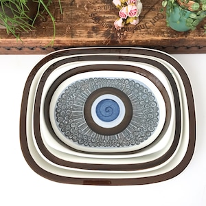 Puede incluir: Juego de tres platos de servir ovalados de cerámica encajables con borde marrón y interior blanco. El plato central presenta un diseño en forma de ojo azul y marrón con un patrón de espiral.