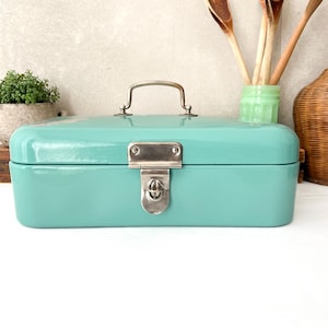 Op de afbeelding: Een rechthoekige, turquoise broodtrommel met een zilveren handvat en sluiting. De broodtrommel heeft afgeronde randen en een glanzende afwerking. De handgreep is gebogen en de sluiting is in het midden geplaatst. Het is een keukenopbergartikel.