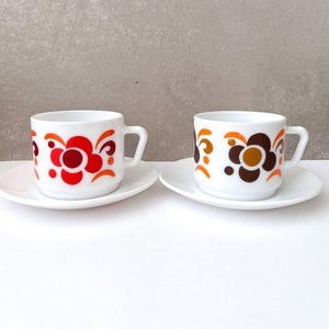 Puede incluir: Dos tazas de té de cerámica blanca con platillos. Las tazas tienen un diseño floral retro en naranja, marrón y rojo.