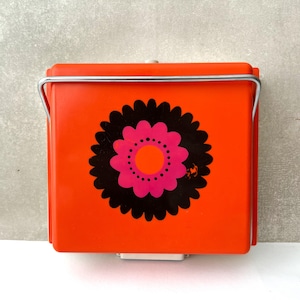May include: A square, orange container with a black and pink floral design. The container has a silver handle and a white base. The floral design is centered on the front of the container. The container is likely vintage or retro.