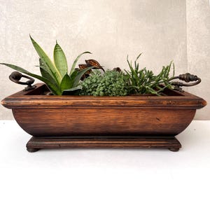 以下が含まれることがあります： 濃い茶色の仕上げが施された長方形の木製プランターで、さまざまな緑色の植物が植えられています。 プランターの両端には装飾的な金属製のハンドルが付いています。 植物には、サンスベリア、小さな緑の葉、その他の緑が含まれています。