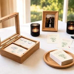 Op de afbeelding: Een houten doos met een scharnierend glazen deksel bevat kaarten met blauwe bloemmotieven en de tekst Lake & Carrie. Daarnaast liggen stapels kaarten met groene bloemillustraties en de tekst Ryan & Anna, een houten dienblad, twee zwarte kaarsenhouders en een fotolijst.