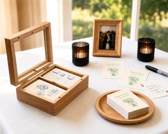Puede incluir: Una caja de madera con tapa de cristal abatible que contiene tarjetas blancas con diseños florales y de texto en azul. Cerca hay pilas de tarjetas blancas con estampados florales verdes, un marco de fotos de madera, dos portavelas negros y un rotulador negro.