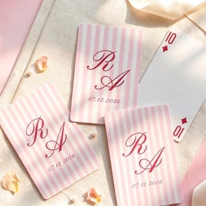 Puede incluir: Cartas de juego a rayas rosas y blancas, con las iniciales "R A" en escritura burdeos y la fecha "07.12.2026". Una carta muestra el número 10 de diamantes. Las cartas están dispuestas sobre una superficie clara con pétalos dispersos.