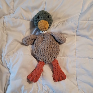 May include: A crocheted plush toy duck with a green head, yellow beak, gray body, and orange legs. The duck is lying on a white blanket.