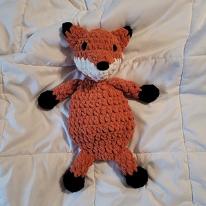 May include: A crocheted fox plush toy with orange fur, black eyes, a white nose, and black feet. The toy is lying on a white quilted blanket.