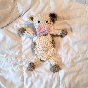 May include: A handmade crochet cow plushie in cream, brown, and pink. The cow has a cream-colored body with brown spots, a pink muzzle, and brown ears and hooves. The plushie is lying on a white surface.