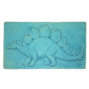 Könnte beinhalten: Eine rechteckige, türkisfarbene Fliese mit einem Relief eines Stegosaurus. Der Dinosaurier ist im Profil dargestellt, mit seinen Platten und Schwanzspitzen. Die Fliese hat eine strukturierte Oberfläche. Die Maße sind in Zentimetern.