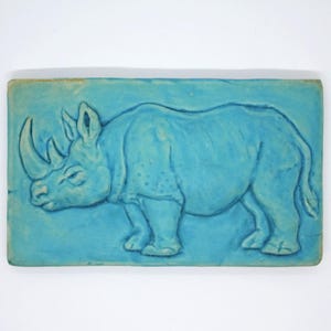 Könnte beinhalten: Türkisfarbene Reliefskulptur eines Nashorns. Das Nashorn ist im Profil dargestellt, mit detaillierten Merkmalen wie Horn, Ohren und strukturierter Haut. Das Kunstwerk ist auf einer rechteckigen Basis montiert.