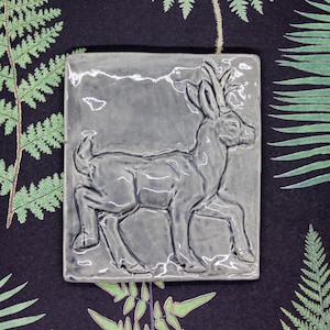 Könnte beinhalten: Rechteckige, graue Keramikfliese mit einem Relief eines Hirsches. Der Hirsch ist im Profil dargestellt, mit detailliertem Geweih und Beinen. Die Fliese hat eine glänzende Oberfläche und ist vor einem dunklen Hintergrund mit Farnmustern platziert.
